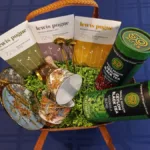 Gift basket on a blue surface containing three Lewis & Poque artisan chocolate bars, two 50 Acres tea canisters, a decorative teacup and saucer, a small cup, and green moss filler.