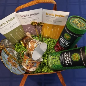 Gift basket on a blue surface containing three Lewis & Poque artisan chocolate bars, two 50 Acres tea canisters, a decorative teacup and saucer, a small cup, and green moss filler.