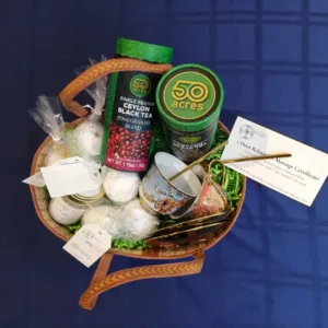 Gift basket with 50 Acres Ceylon Black Tea, a green tea tin, a mug, wrapped treats, and a massage certificate on a blue table.