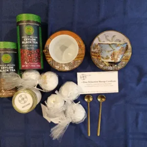 Assorted tea cans, two decorative teacups with saucers, wrapped bath items, a '1-Hour Relaxation Massage' certificate, a small 'Recover' tin, and two gold spoons on a blue cloth.