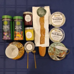 Flat lay of tea tins, jams, a baguette in a white bag, and labeled spreads with two decorative teacups on a blue tablecloth (breakfast/tea setup).