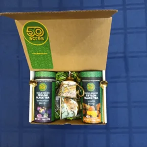 Open gift box showing two 50 Acres Ceylon Black Tea bottles (Mango Blend) and a decorative teapot centerpiece on a blue surface